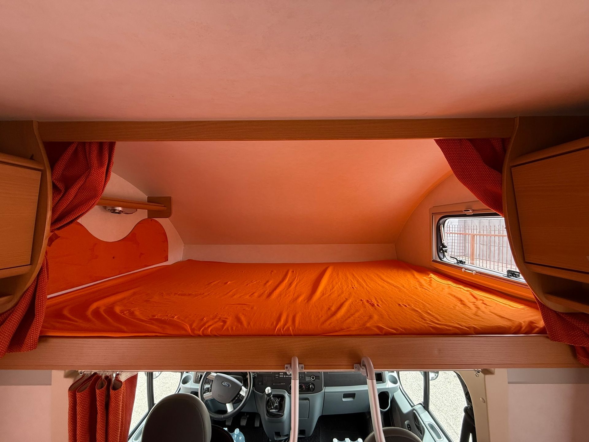 Zona notte sopraelevata in un camper con letto arancione e tende. La parte anteriore del camper è visibile sotto.