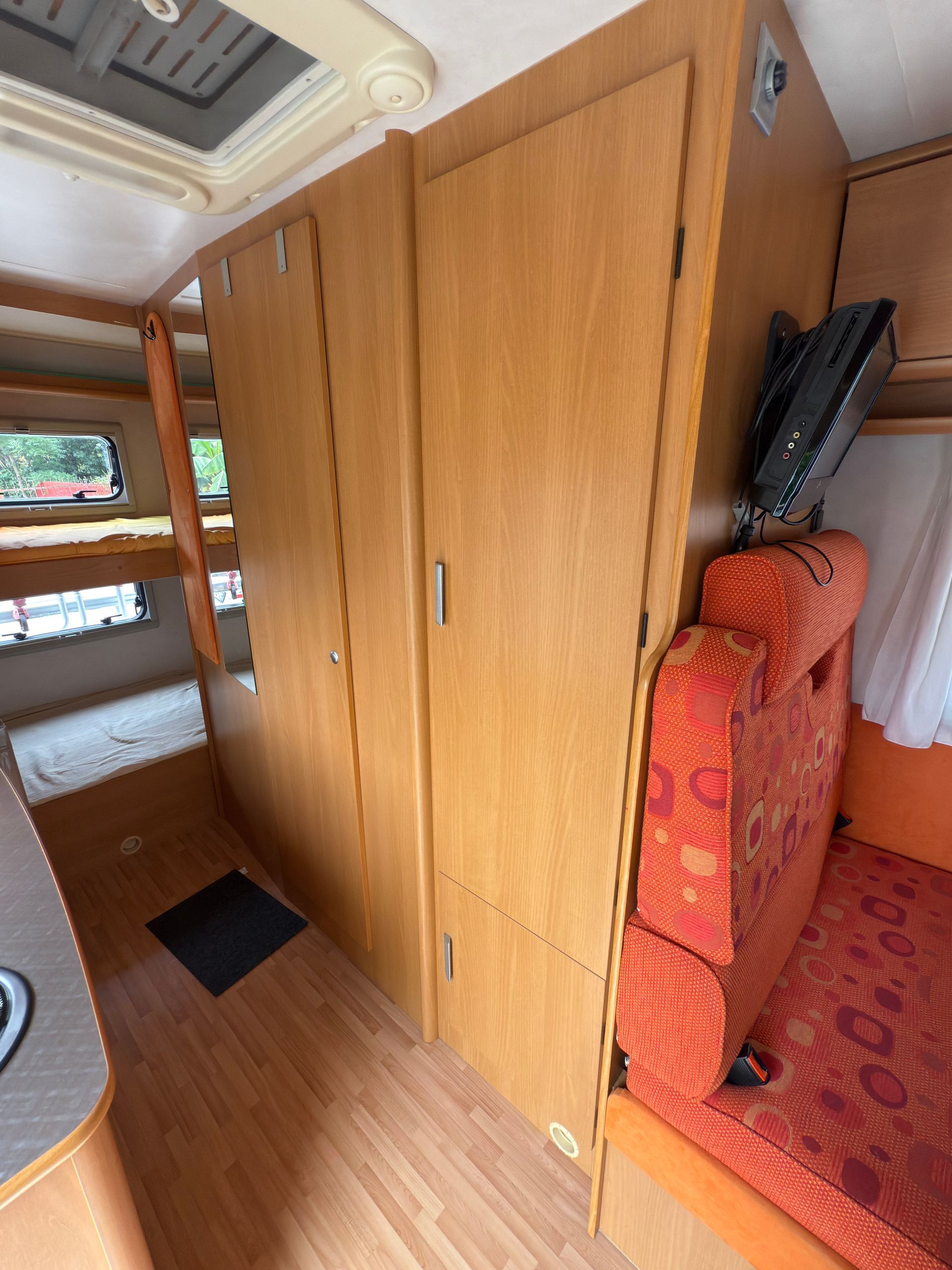Interno di un camper con mobili in legno, sedili arancioni e un letto a castello visibile sullo sfondo.