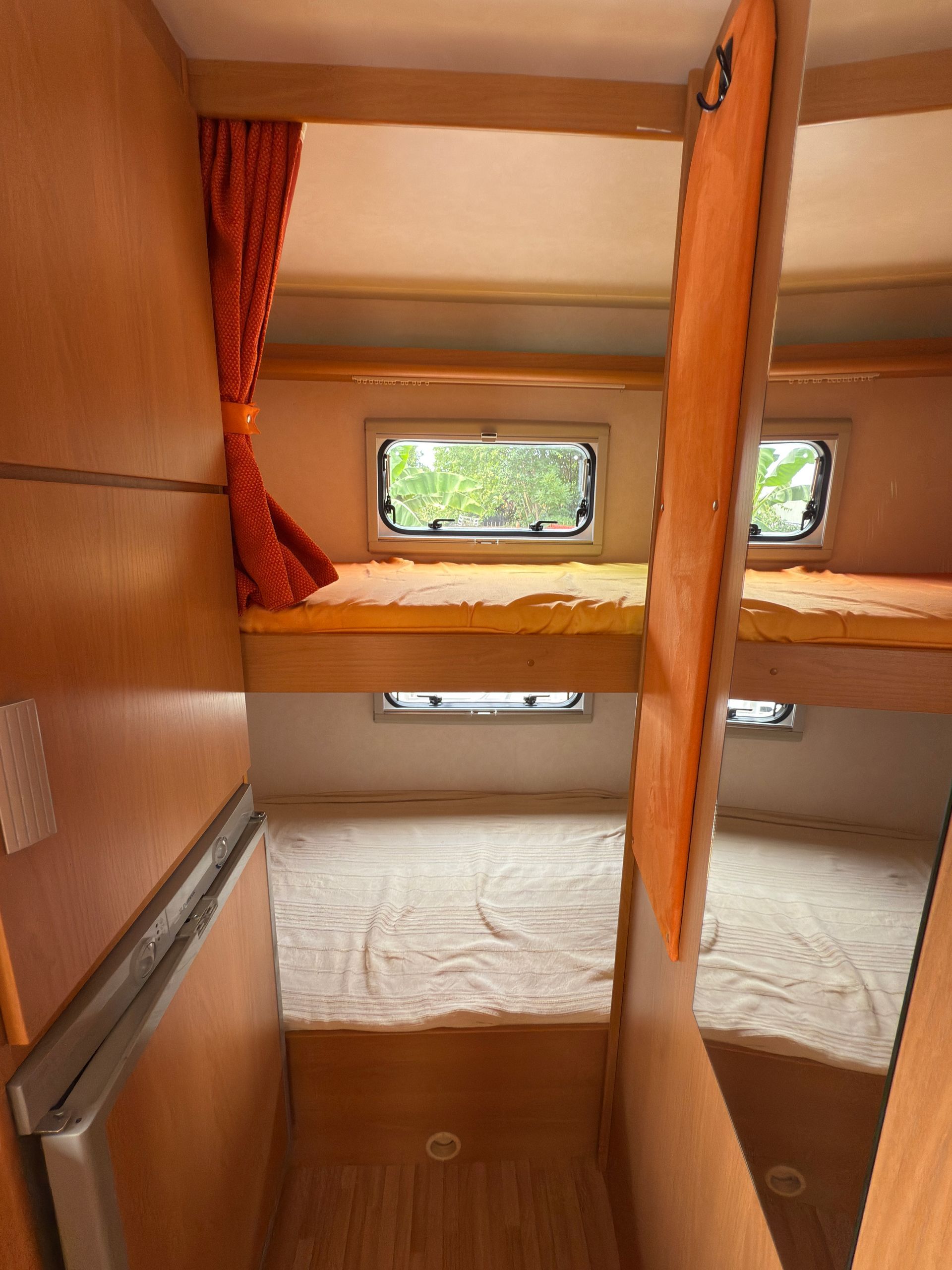 Interno di un piccolo camper con letti a castello, finestre e tende arancioni.