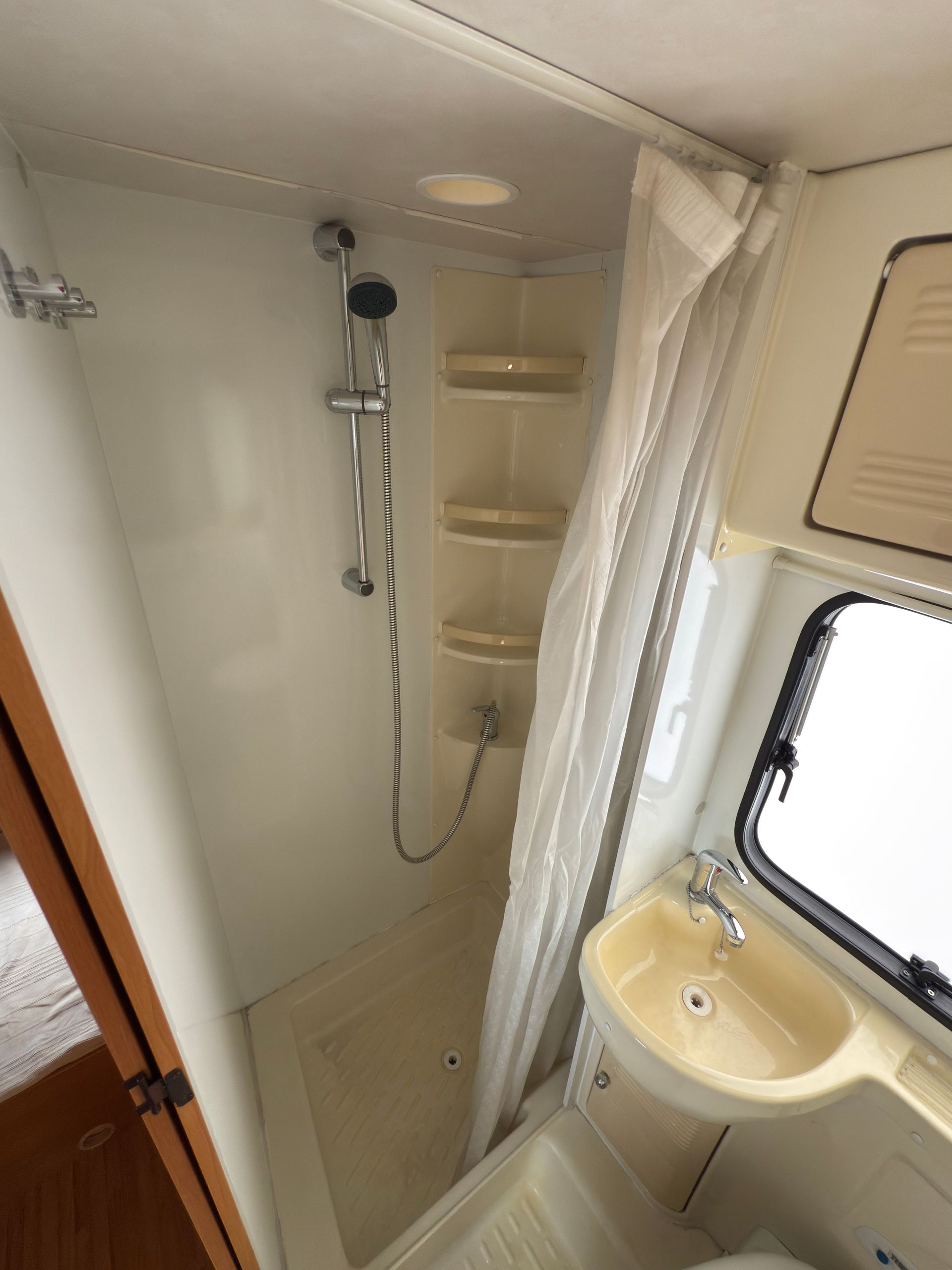Piccolo bagno per camper con doccia, lavandino e ripiani; bianco e beige.