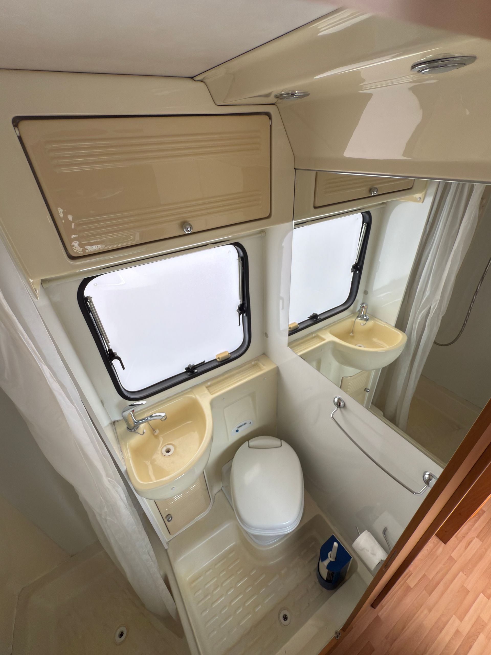 Piccolo bagno per camper con WC, lavandini, specchio, finestra e mobiletto. Interni beige e bianchi.