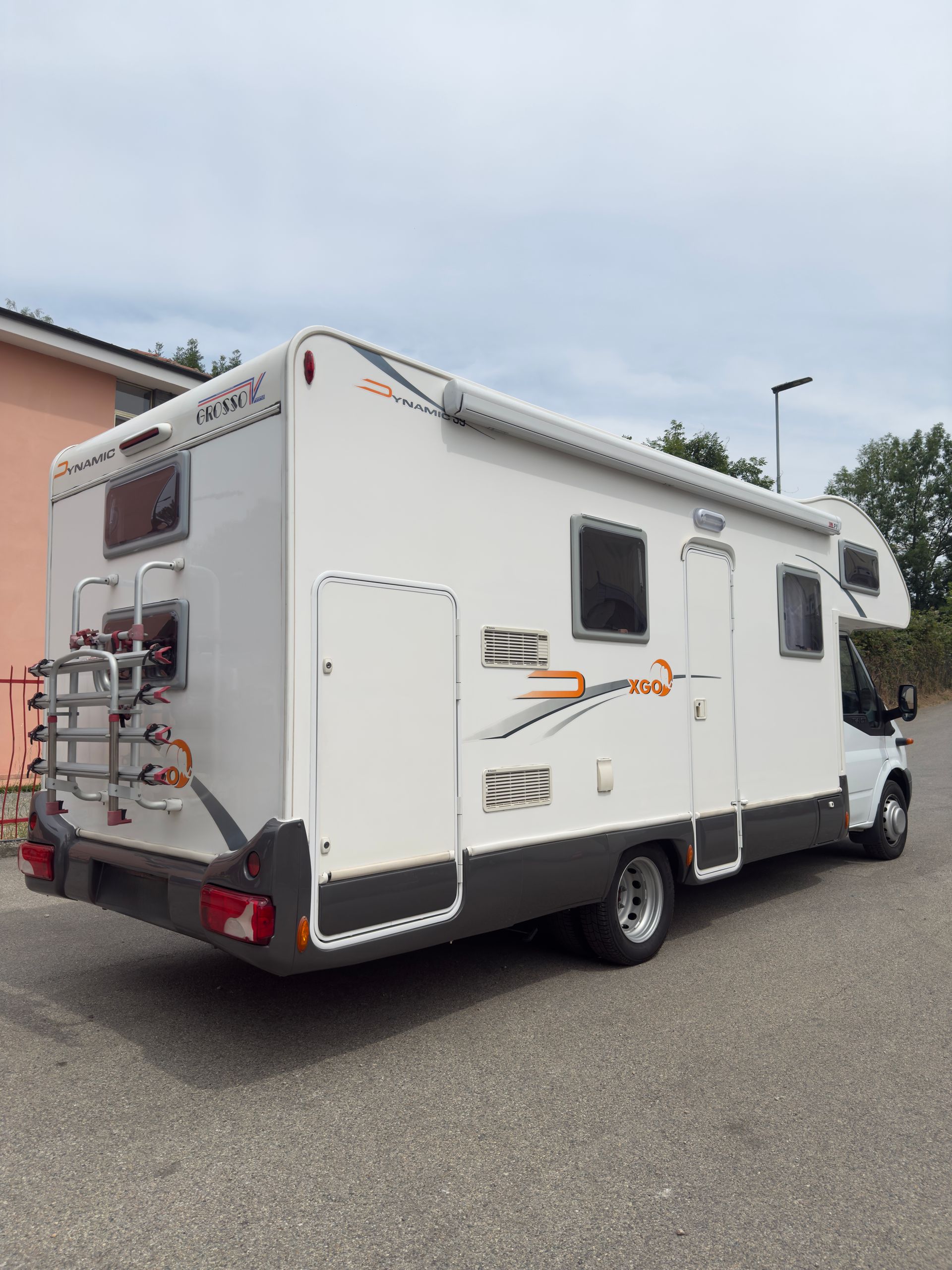 Veicolo ricreativo bianco parcheggiato sul marciapiede. Dotato di portabici posteriore e tendalino.