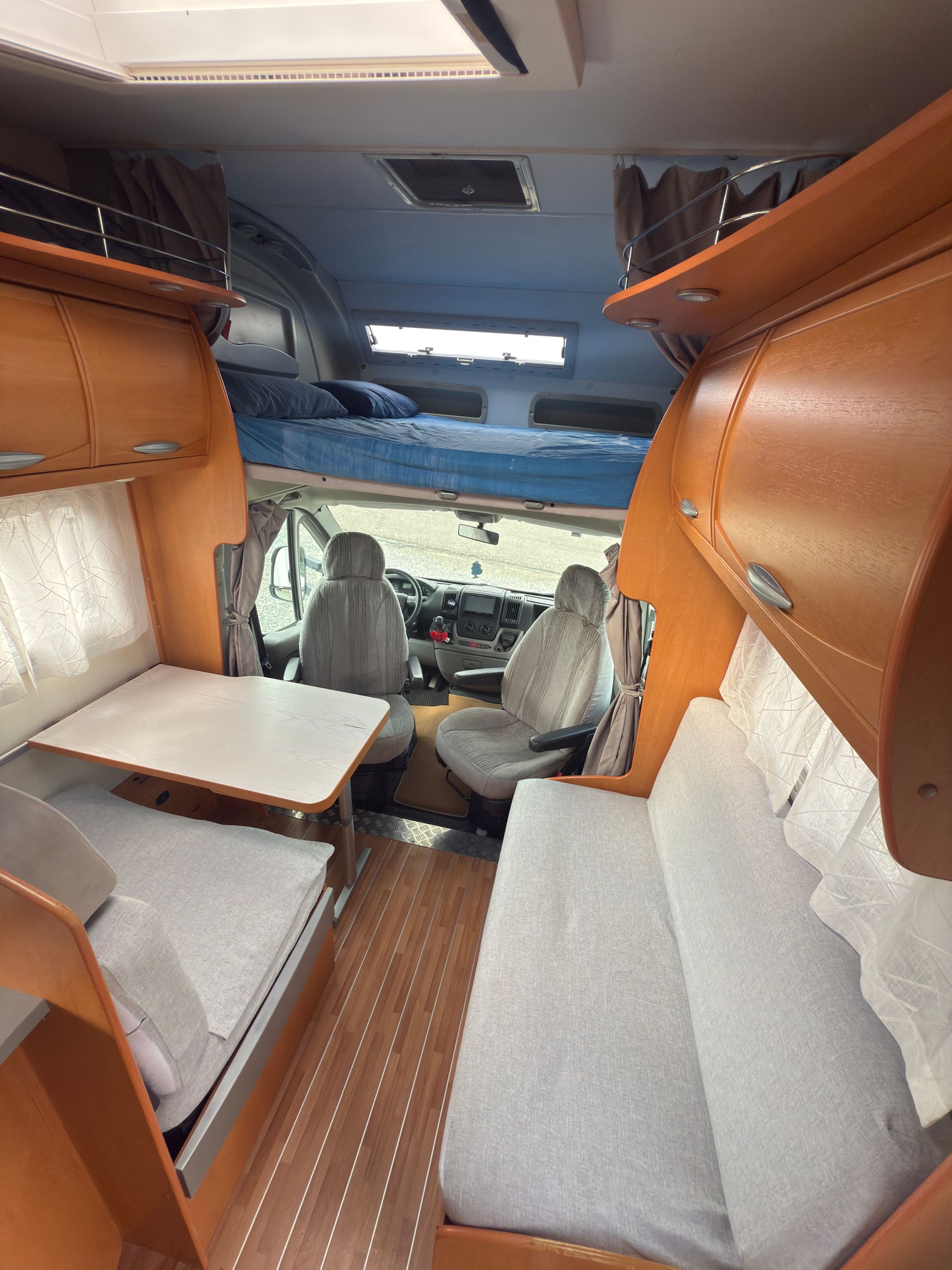 Interno di un camper con posti a sedere, tavolo, vani portaoggetti superiori e postazione di guida visibili in lontananza.