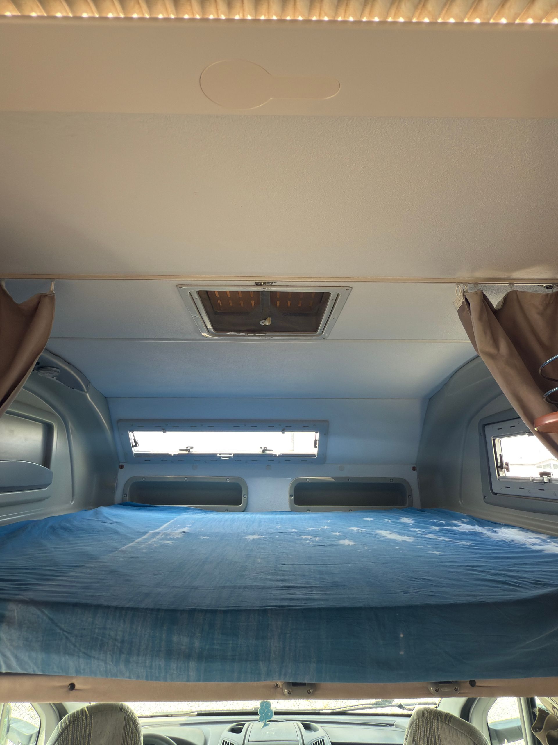 Vista interna di un camper con un letto, piccole finestre e una presa d'aria sul soffitto.
