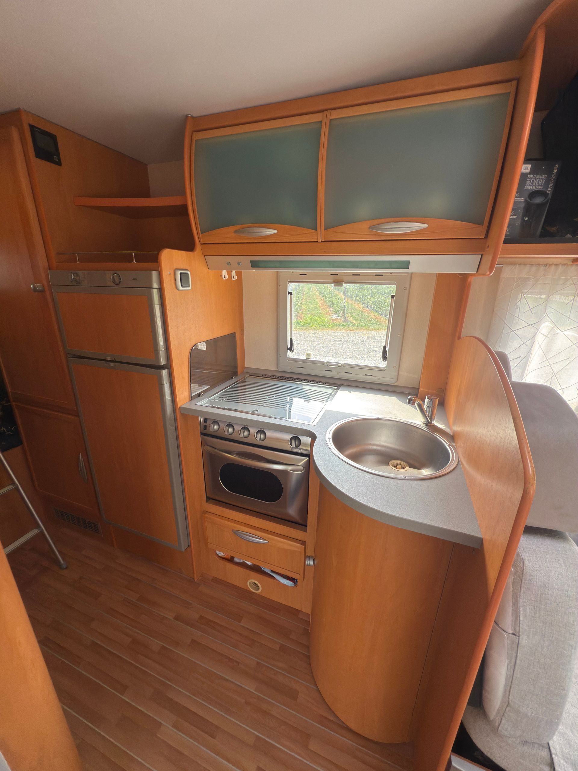 Interno della cucina di un camper con mobili in legno, frigorifero, fornello, forno, lavello e finestra.