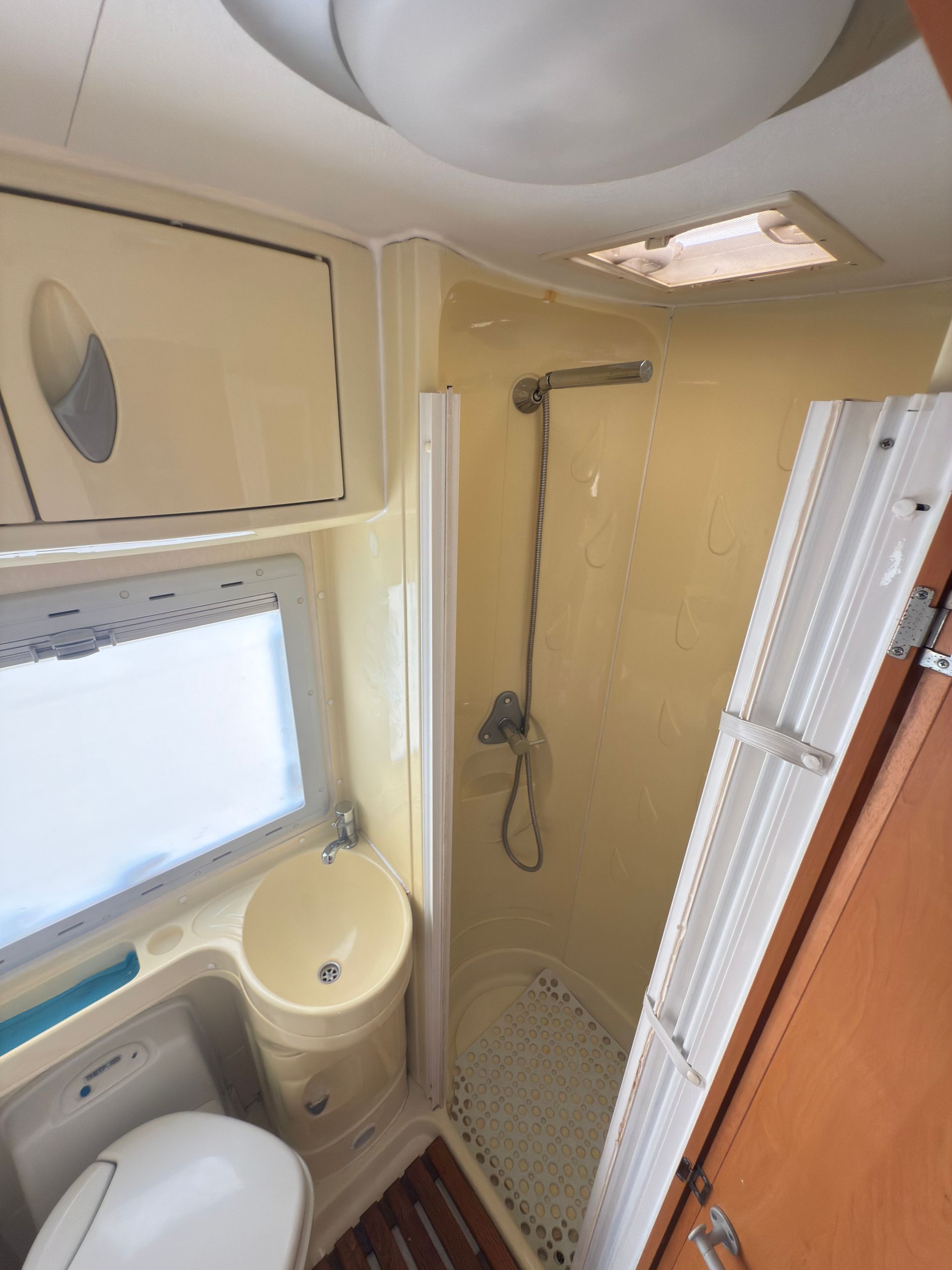 Bagno per camper con doccia, WC, lavandino e piccola finestra. Interni beige e bianchi con pavimento effetto legno.