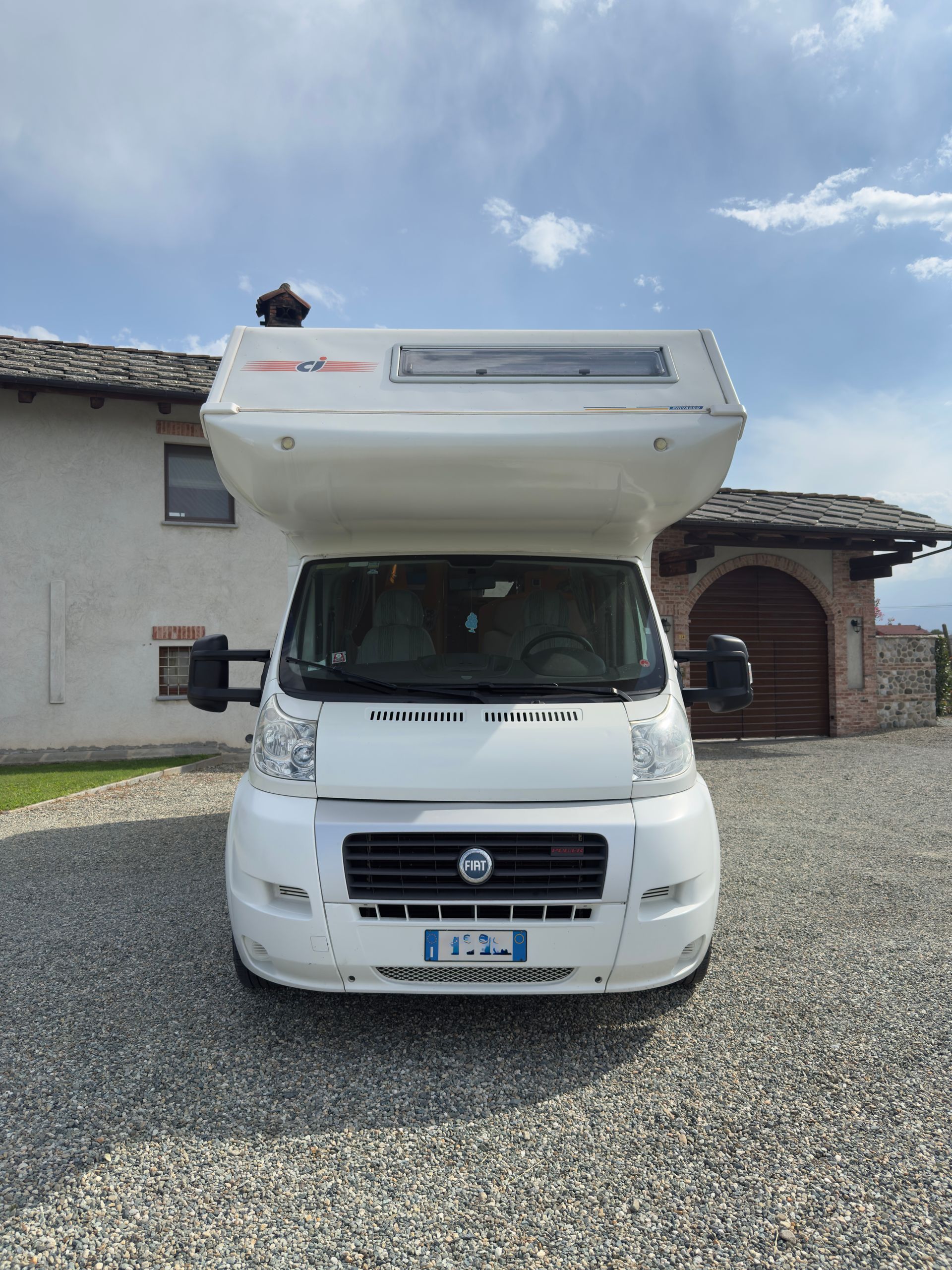 Camper bianco parcheggiato sulla ghiaia di fronte a un edificio con un arco in pietra sotto un cielo nuvoloso.