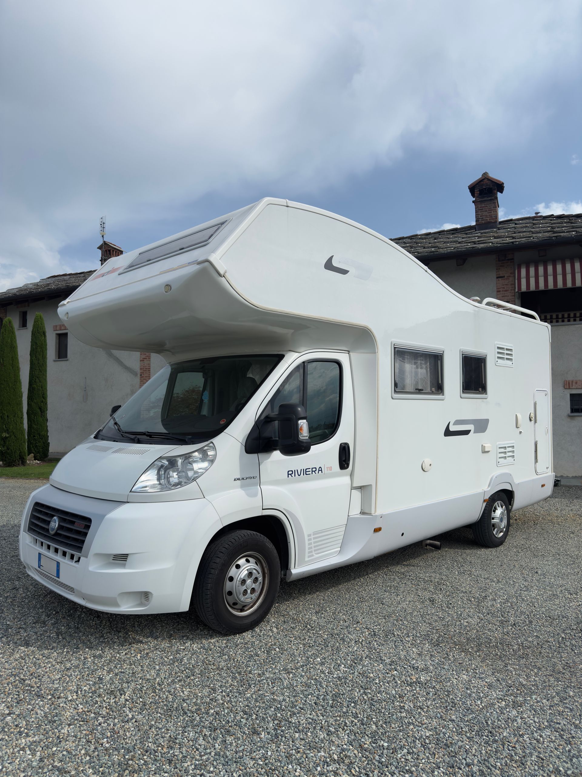 Camper bianco parcheggiato sulla ghiaia con un edificio residenziale sullo sfondo. Cielo nuvoloso.