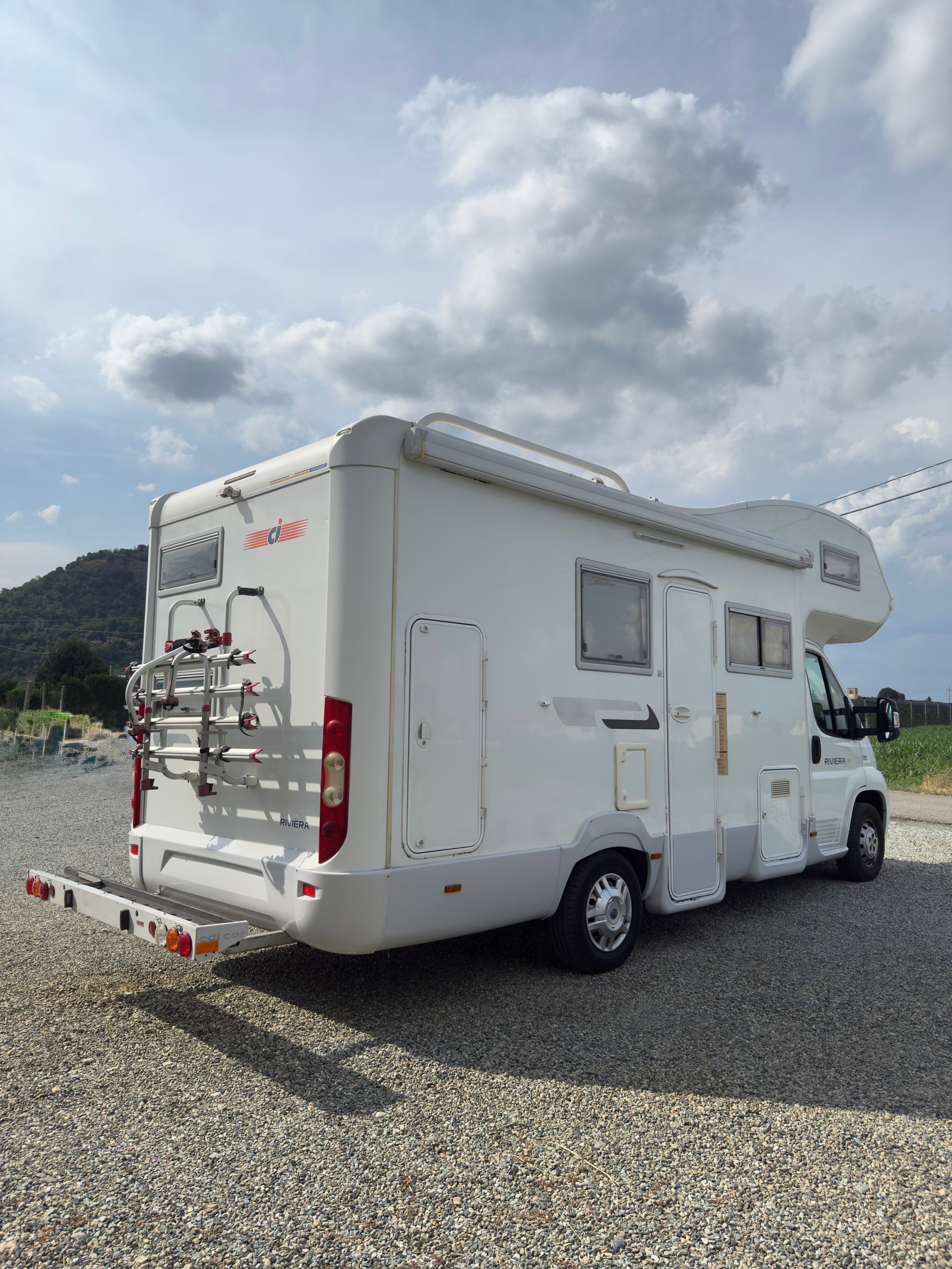 Camper bianco parcheggiato sulla ghiaia. Portabici sul retro, tendalino aperto, cielo nuvoloso.