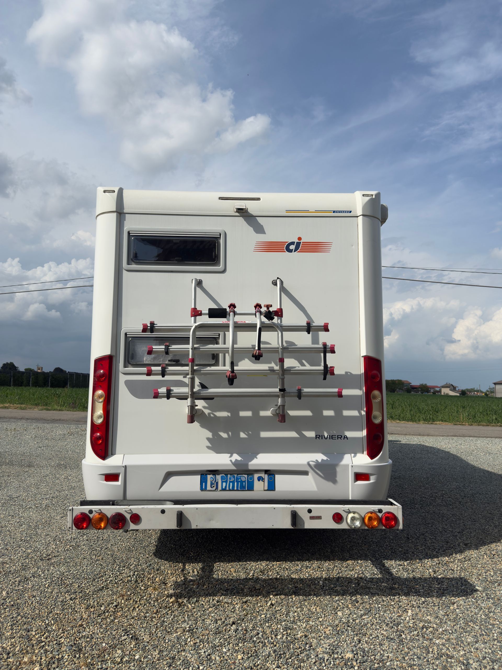 Vista posteriore di un camper bianco con portabiciclette su ghiaia sotto un cielo nuvoloso.