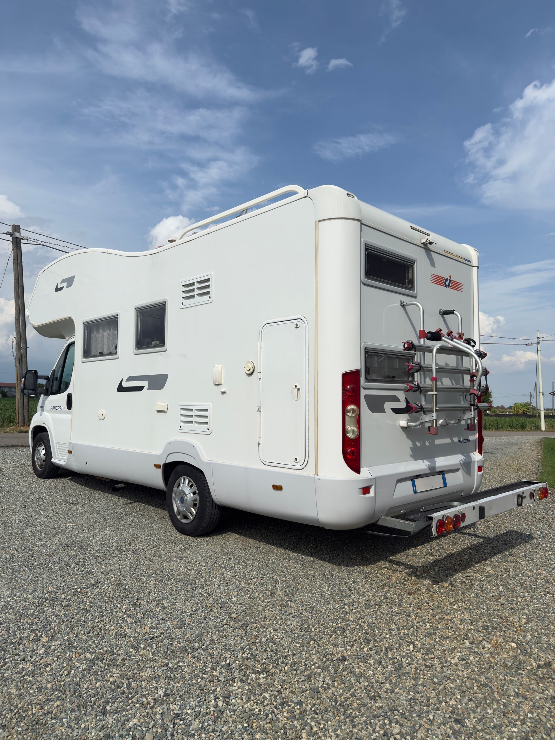 Camper bianco parcheggiato sulla ghiaia sotto un cielo azzurro, con un portabiciclette sul retro.