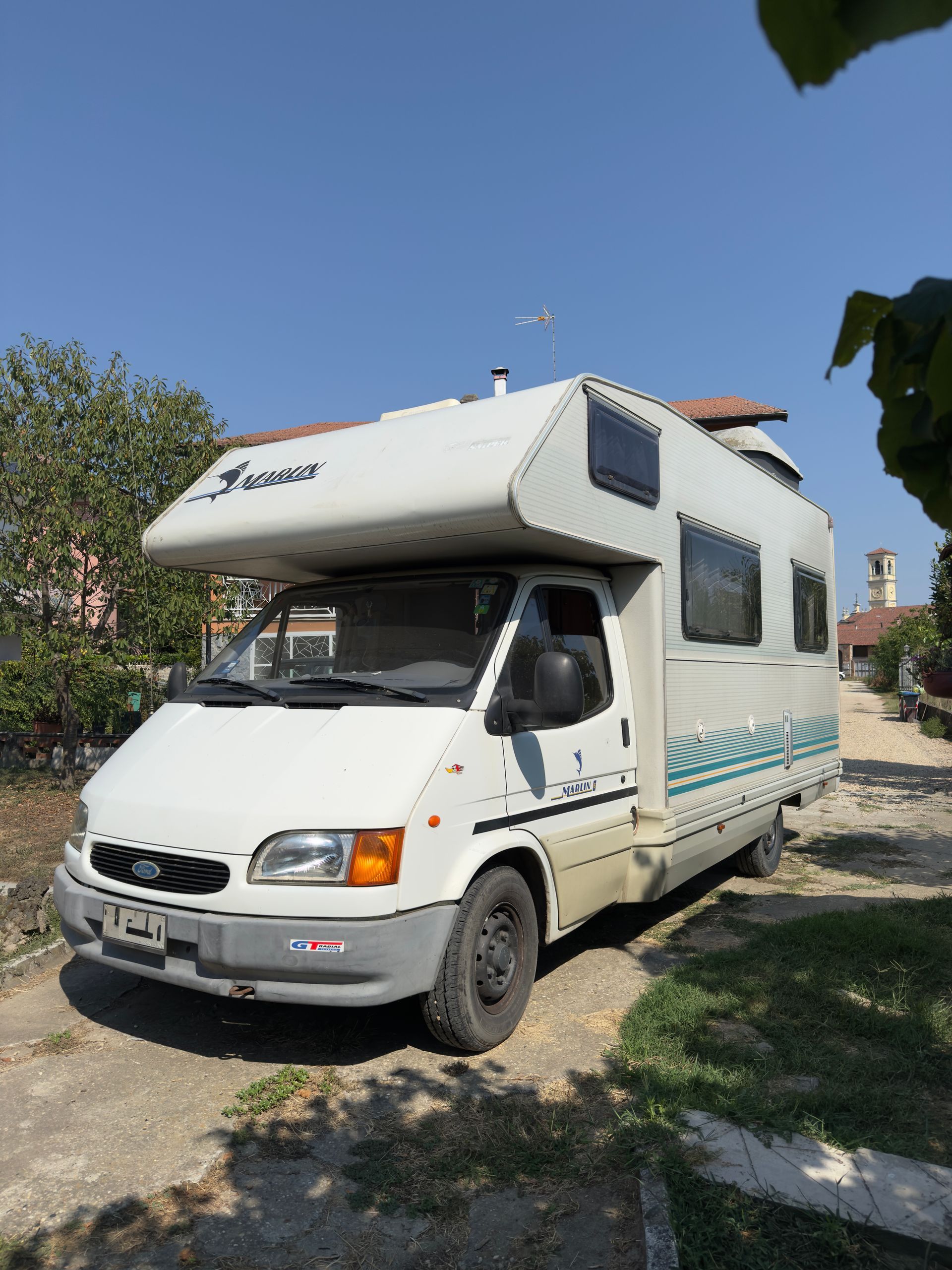 Camper bianco parcheggiato su un vialetto di ghiaia sotto un cielo azzurro e limpido.