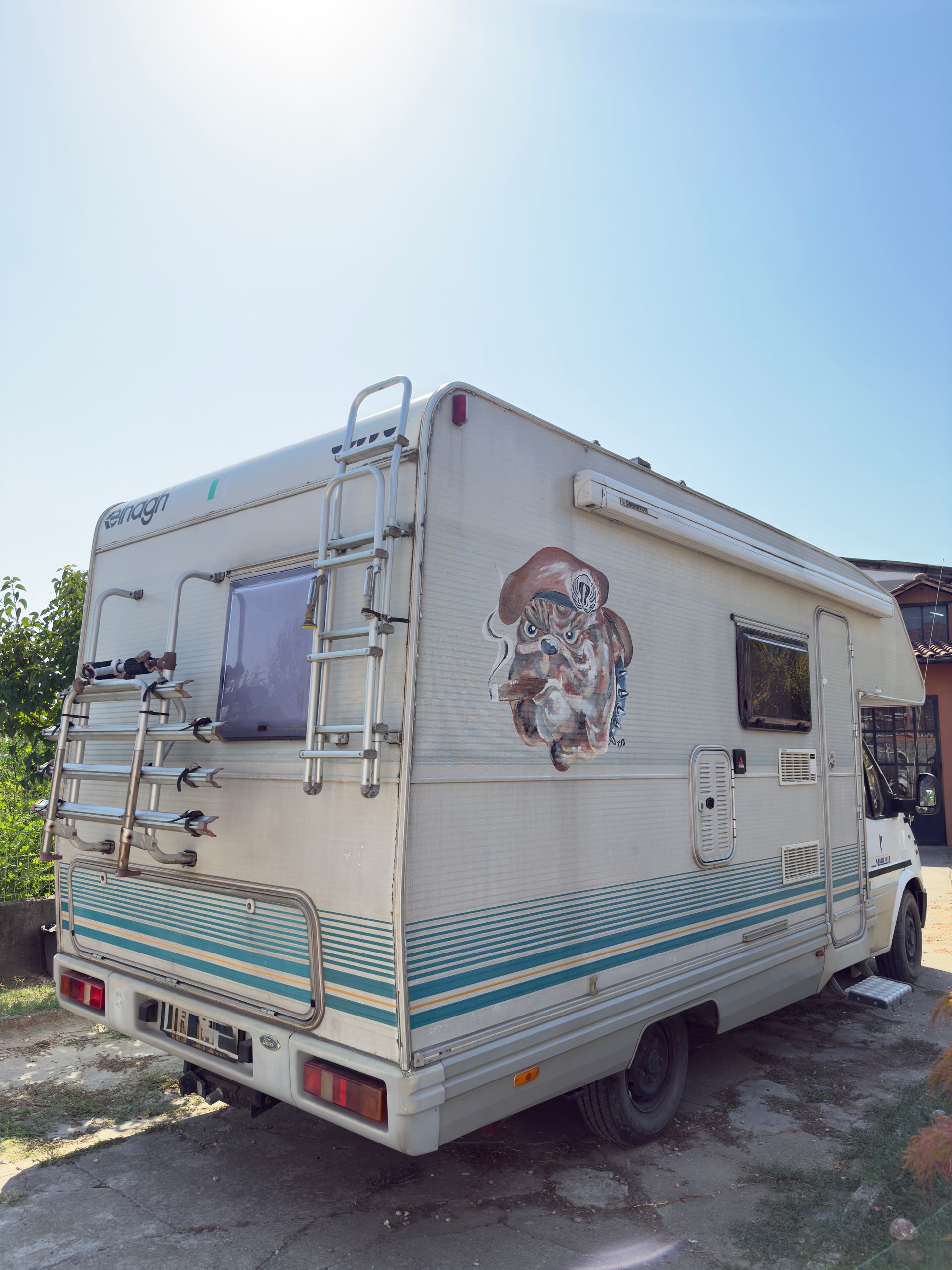 Camper bianco con strisce color verde acqua e scala parcheggiato all'esterno in una giornata di sole.