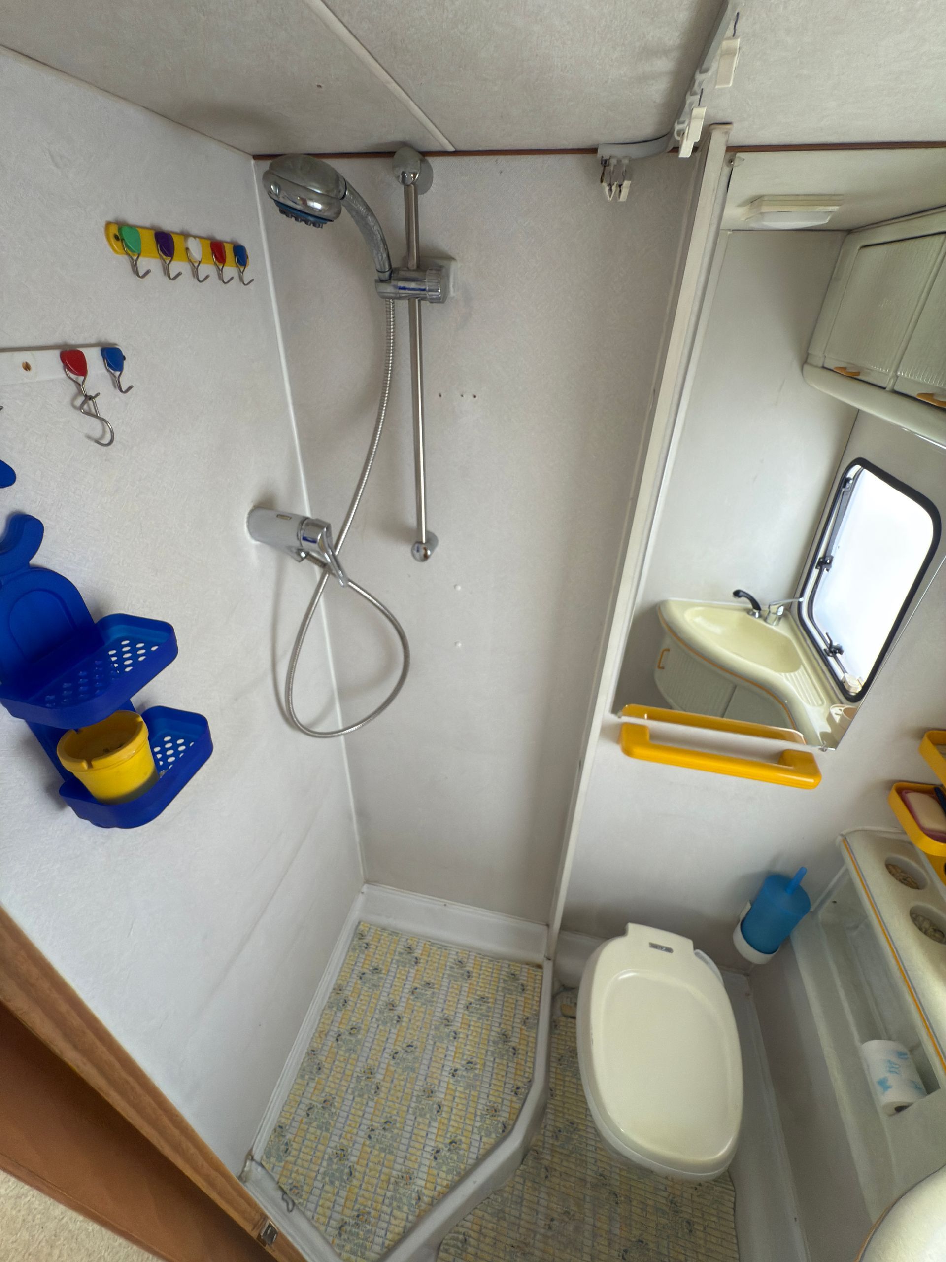 Piccola doccia per imbarcazioni con WC, doccetta e finestra. Pareti bianche, accessori gialli e blu.