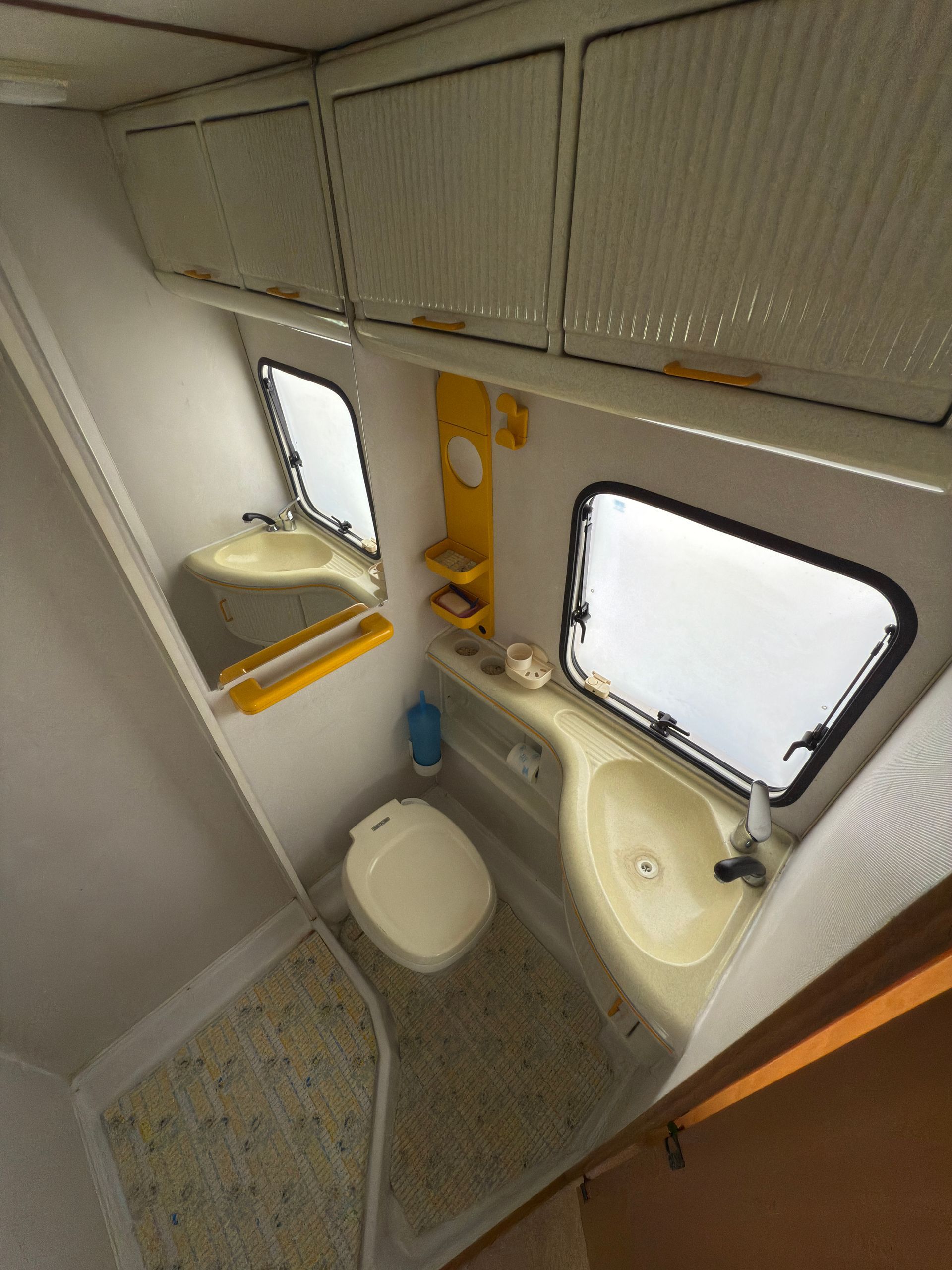 Piccolo bagno per camper con WC, lavandino, specchio e pensili.