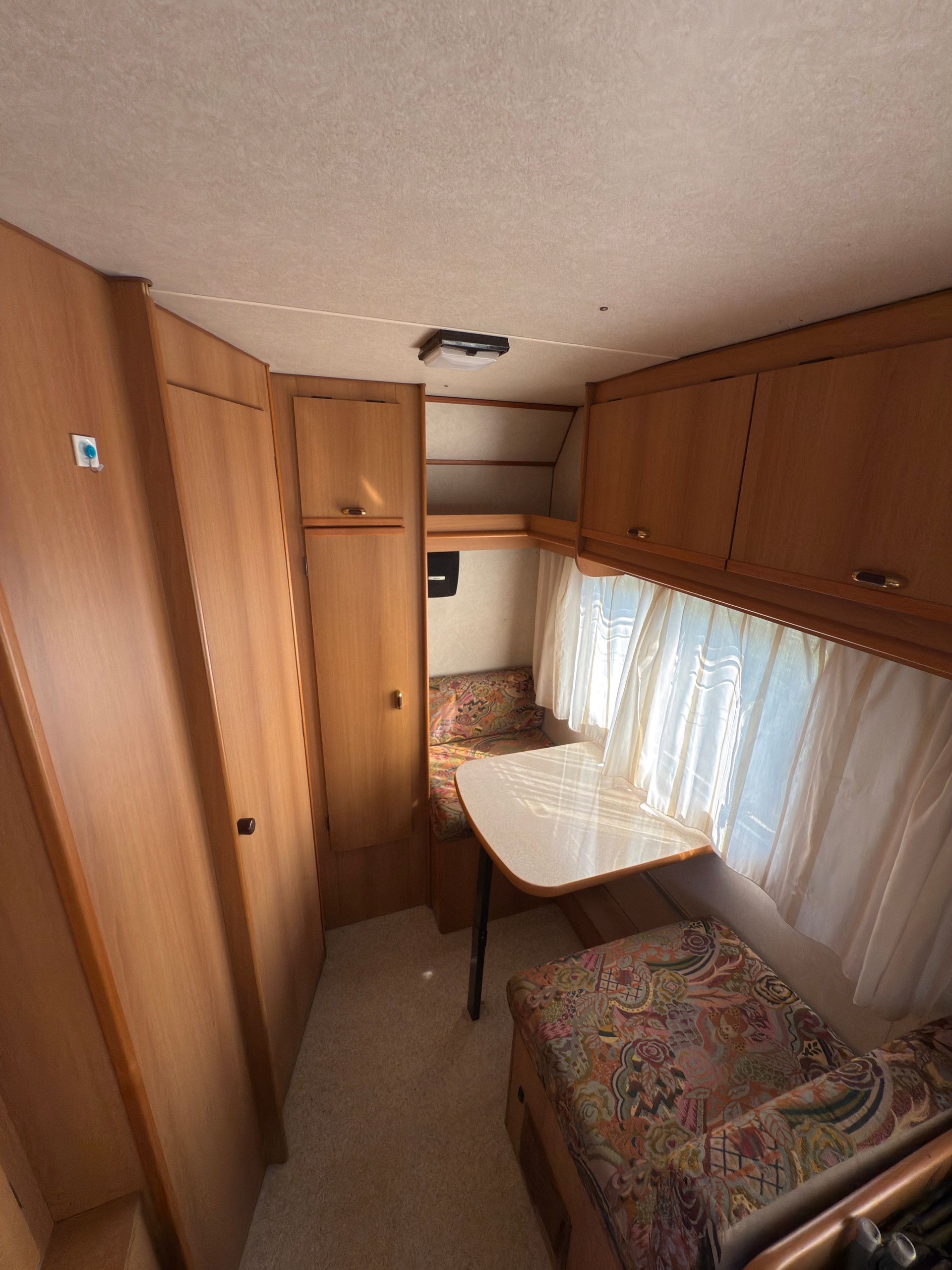 Interno di un camper con mobili in legno, un tavolo e sedute decorate vicino a una finestra con tende.