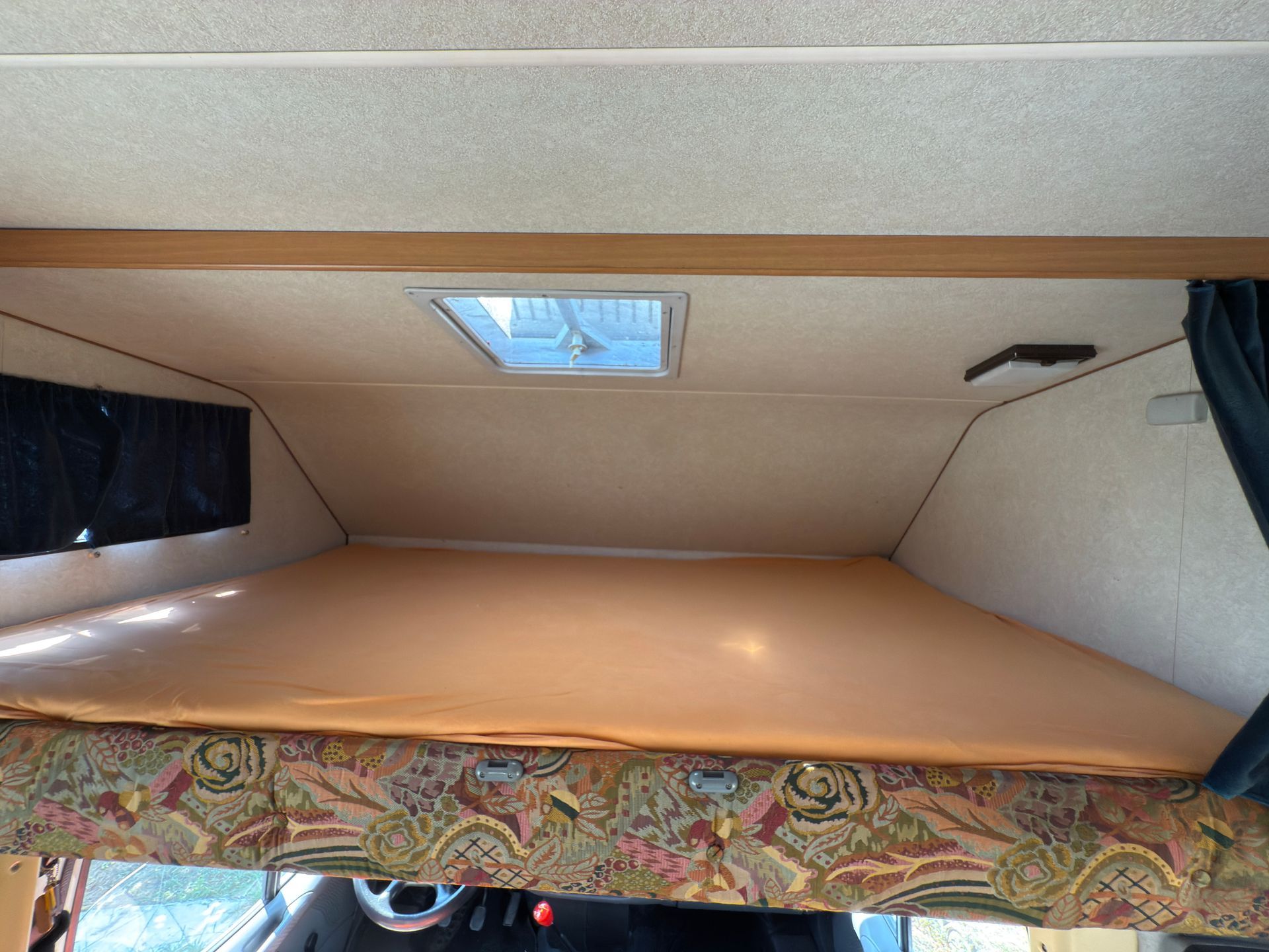 Vista interna di un camper con letto sopraelevato, finestra e tessuto fantasia.