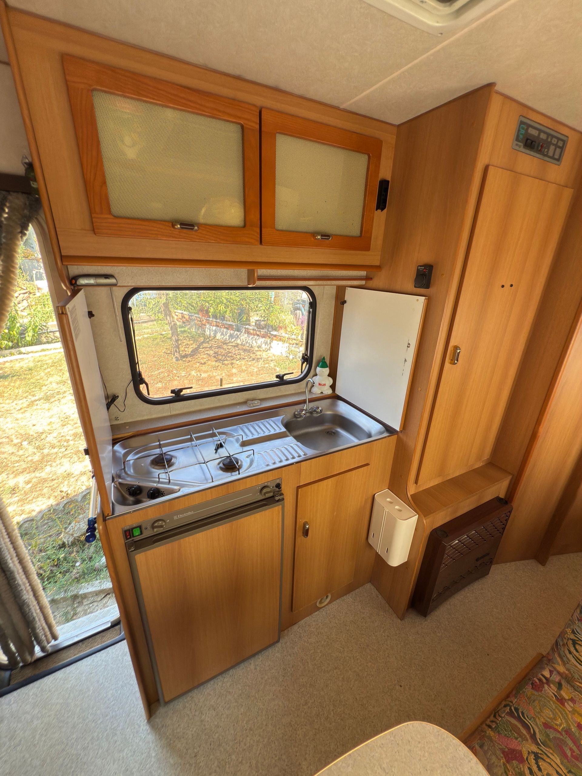 Cucina per camper con mobili, lavello, fornello e frigorifero. Interni in legno con finestra e piccola stufa.