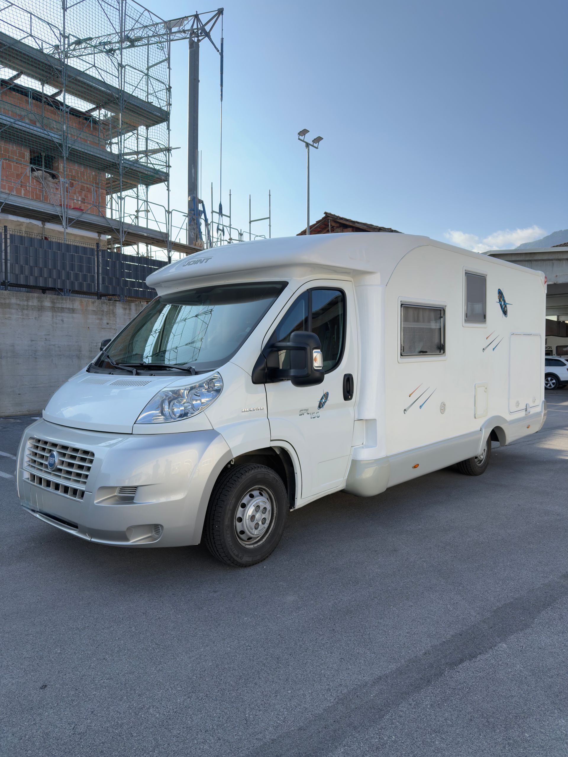 Camper bianco parcheggiato sull'asfalto, con un edificio e il cielo sullo sfondo.