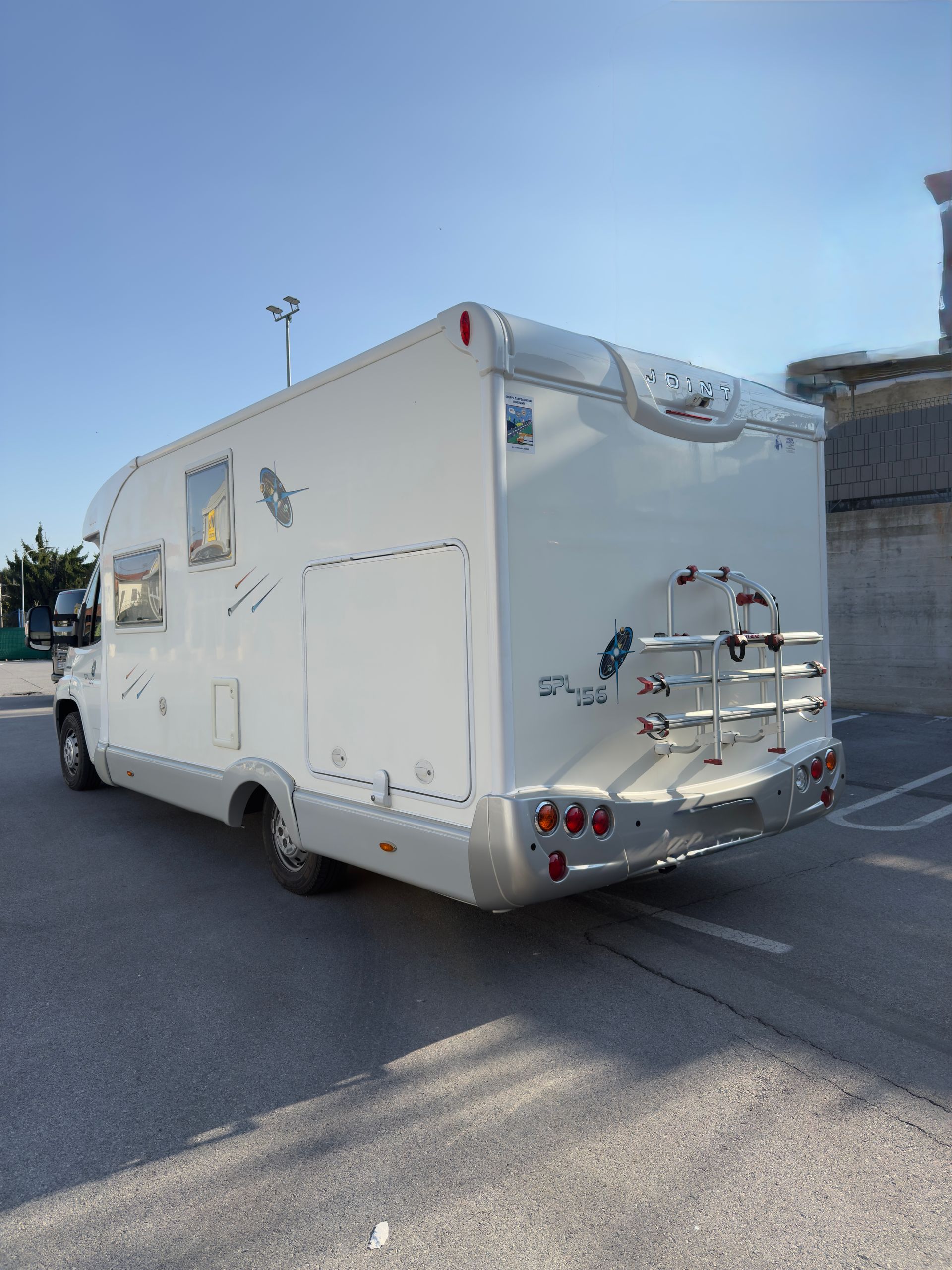 Camper bianco parcheggiato sull'asfalto, vista posteriore con portabici e fanali posteriori sotto il cielo azzurro.
