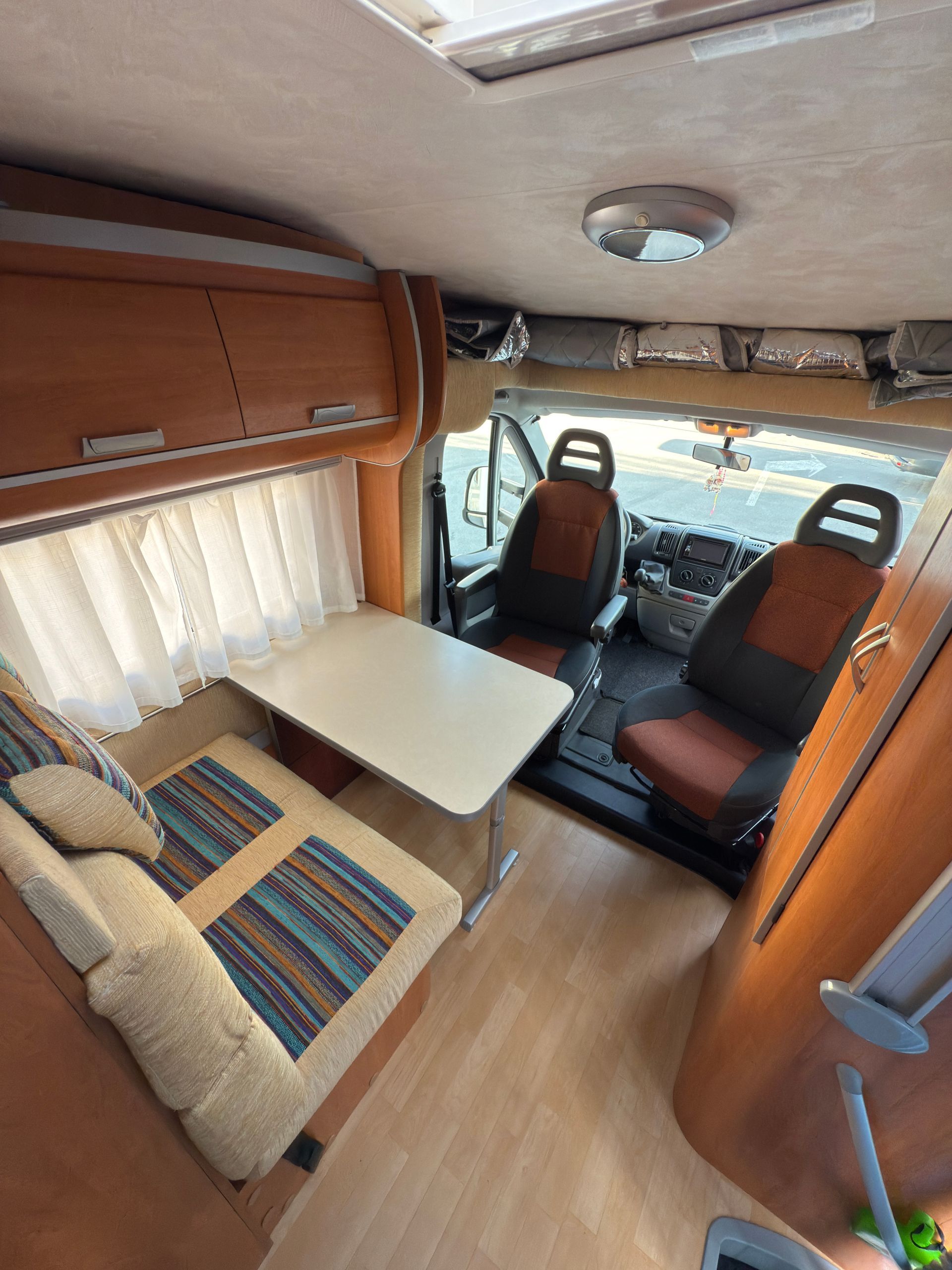 Interno di un camper con area salotto, tavolo e sedili conducente/passeggero. Pannelli in legno chiaro, pavimento chiaro.