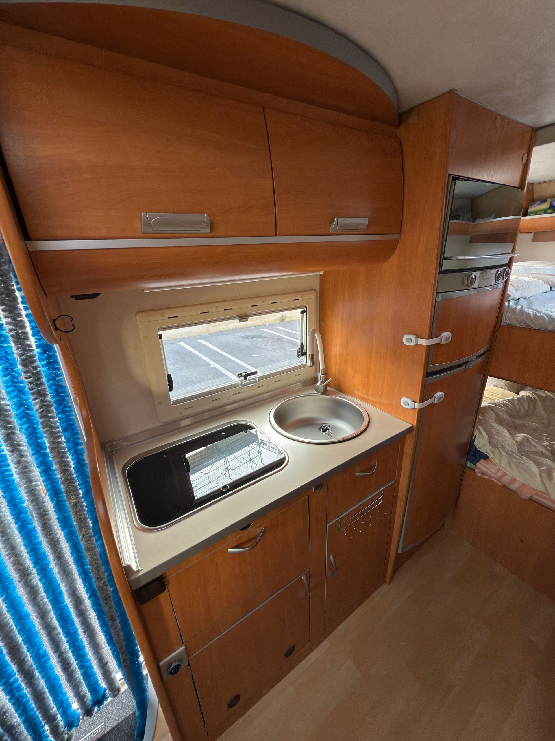 Interno di un camper con mobili in legno, lavandino, fornello e una finestra che si affaccia su un parcheggio.