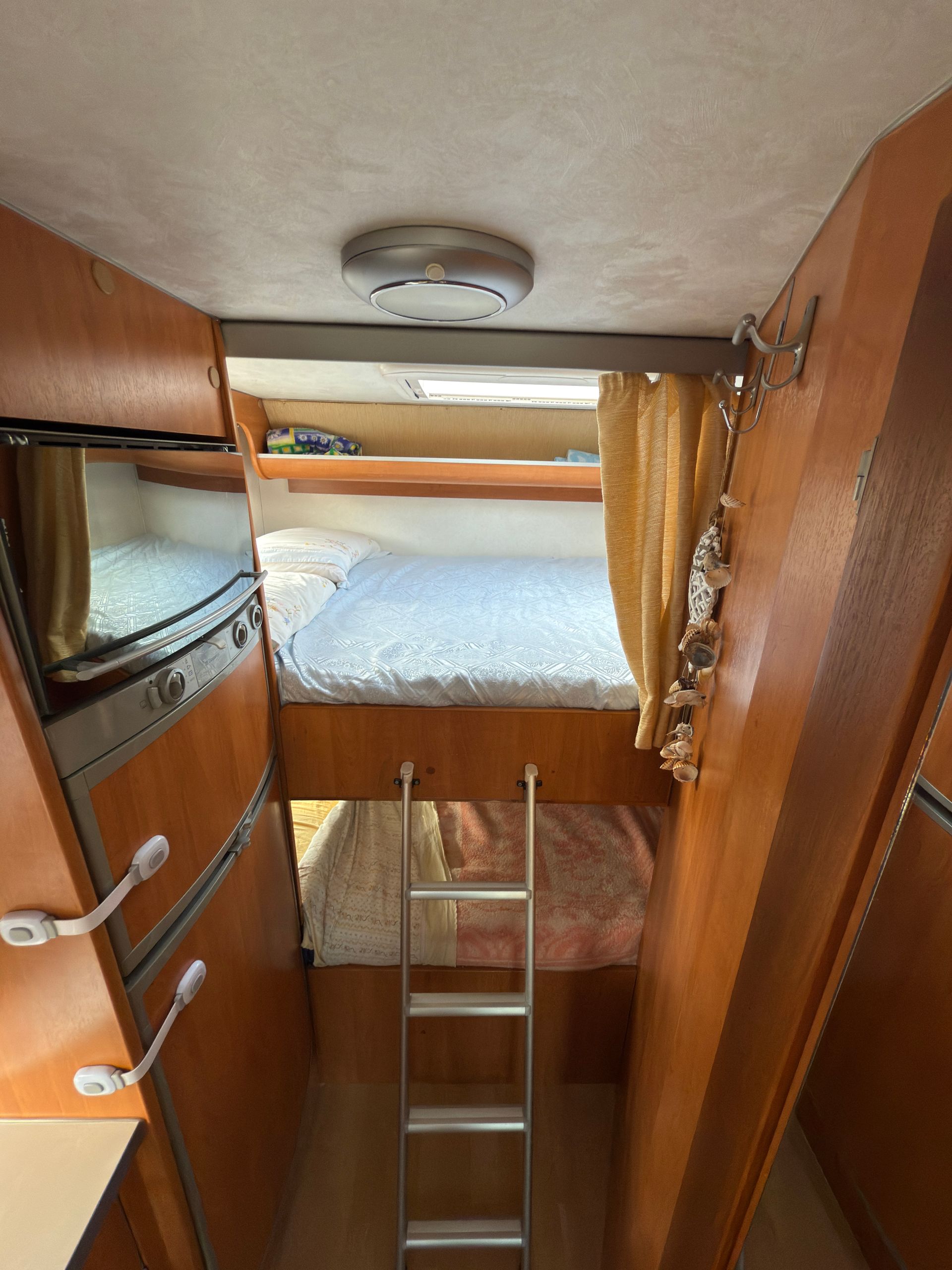 Interno di un camper, con letti a castello, pannelli in legno, una scala e una piccola TV.