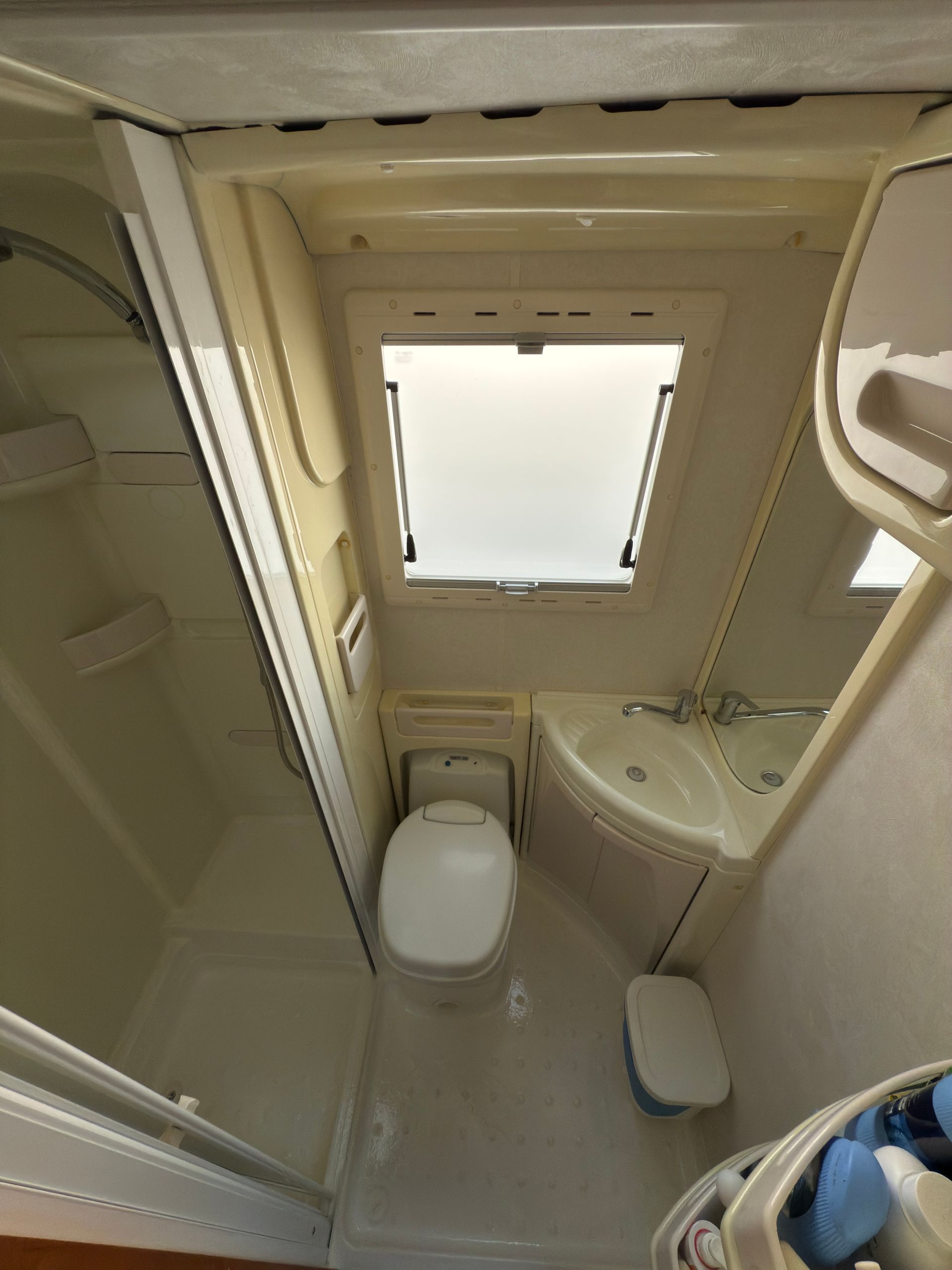 Piccolo bagno per camper con doccia, WC, lavandino e finestra sul soffitto. Pareti e sanitari di colore chiaro.