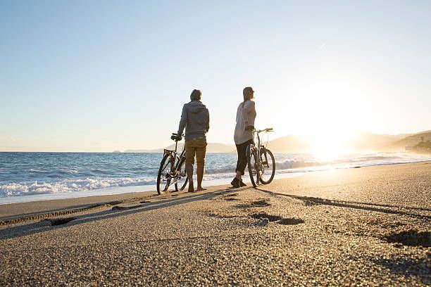 Due persone con le biciclette su una spiaggia, guardano l'oceano al tramonto.