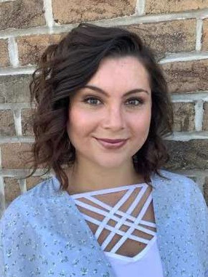 Woman with brown curly hair and makeup smiles in front of a brick wall, wearing a white top with criss-cross straps and a blue floral cardigan.