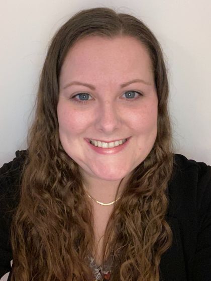 Woman with long brown wavy hair, smiling, wearing a black blazer, gold necklace; neutral background.