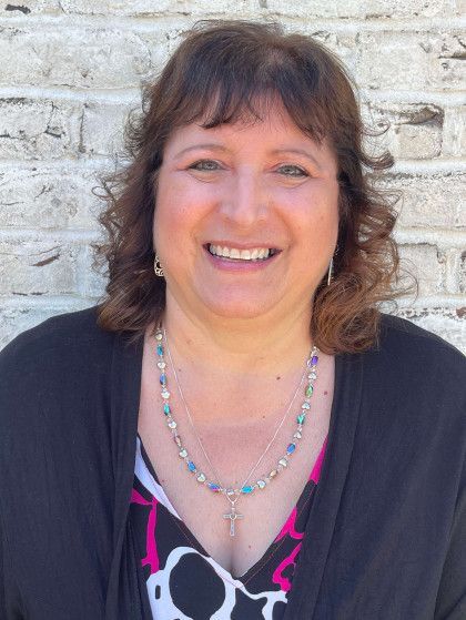 Woman with brown hair, smiling at camera, wearing necklace, standing against brick wall.