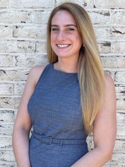 Woman with long blonde hair, wearing a sleeveless blue dress, smiles in front of a brick wall.
