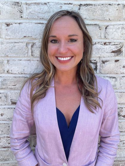 Blonde woman smiles, wearing a pink blazer and blue top, leaning against a brick wall.