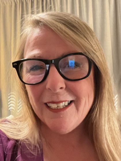 Woman with blonde hair and glasses smiling at the camera, wearing a purple shirt.