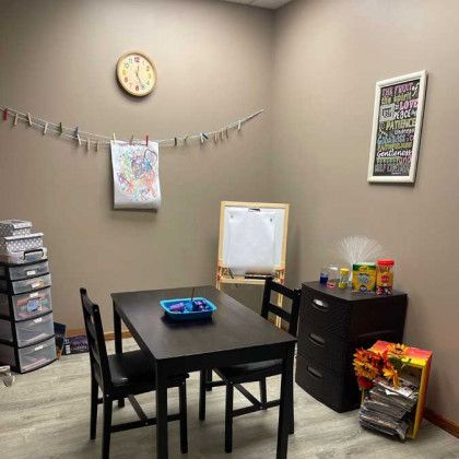 A child's art room: dark table and chairs, art supplies, whiteboard, clock, and a hanging clothesline with artwork.