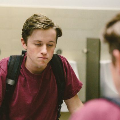 Young man with backpack looking in mirror; maroon shirt, somber expression in bathroom.