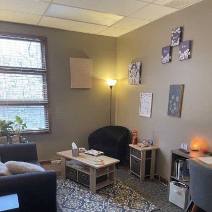 Cozy therapist's office with dark seating, decorative wall art, and a window with blinds.