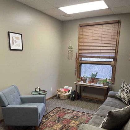 Cozy therapy room: blue chair, gray couch, rug, plants by window.