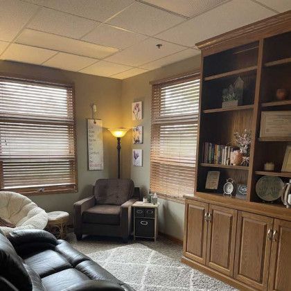 Cozy therapist's office with brown leather sofa, chair, bookshelf, and windows with blinds. Soft lighting.