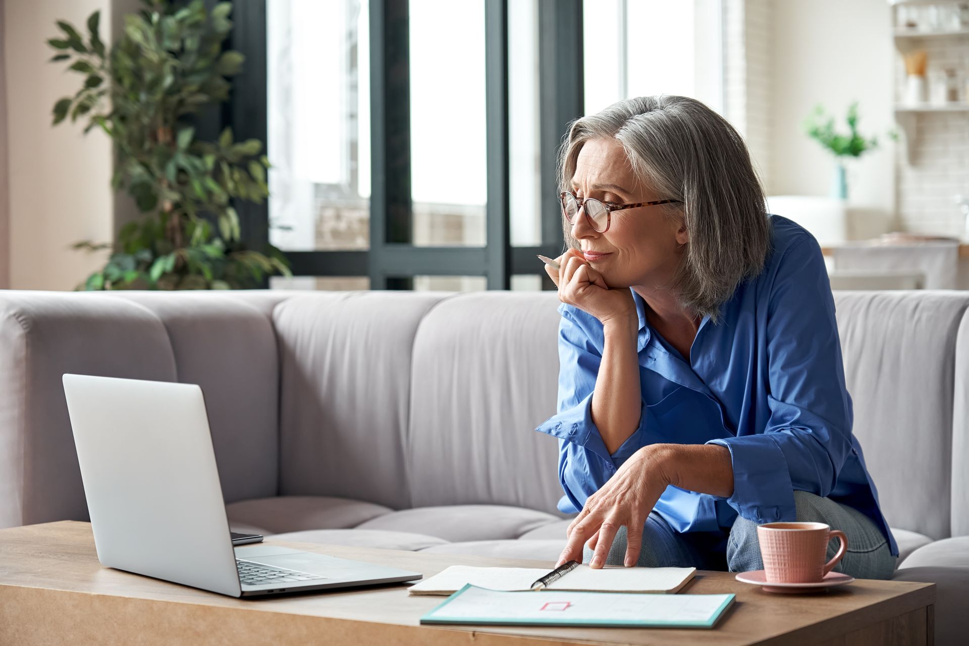 Woman with gray hair and glasses working on laptop on couch, papers and coffee nearby.