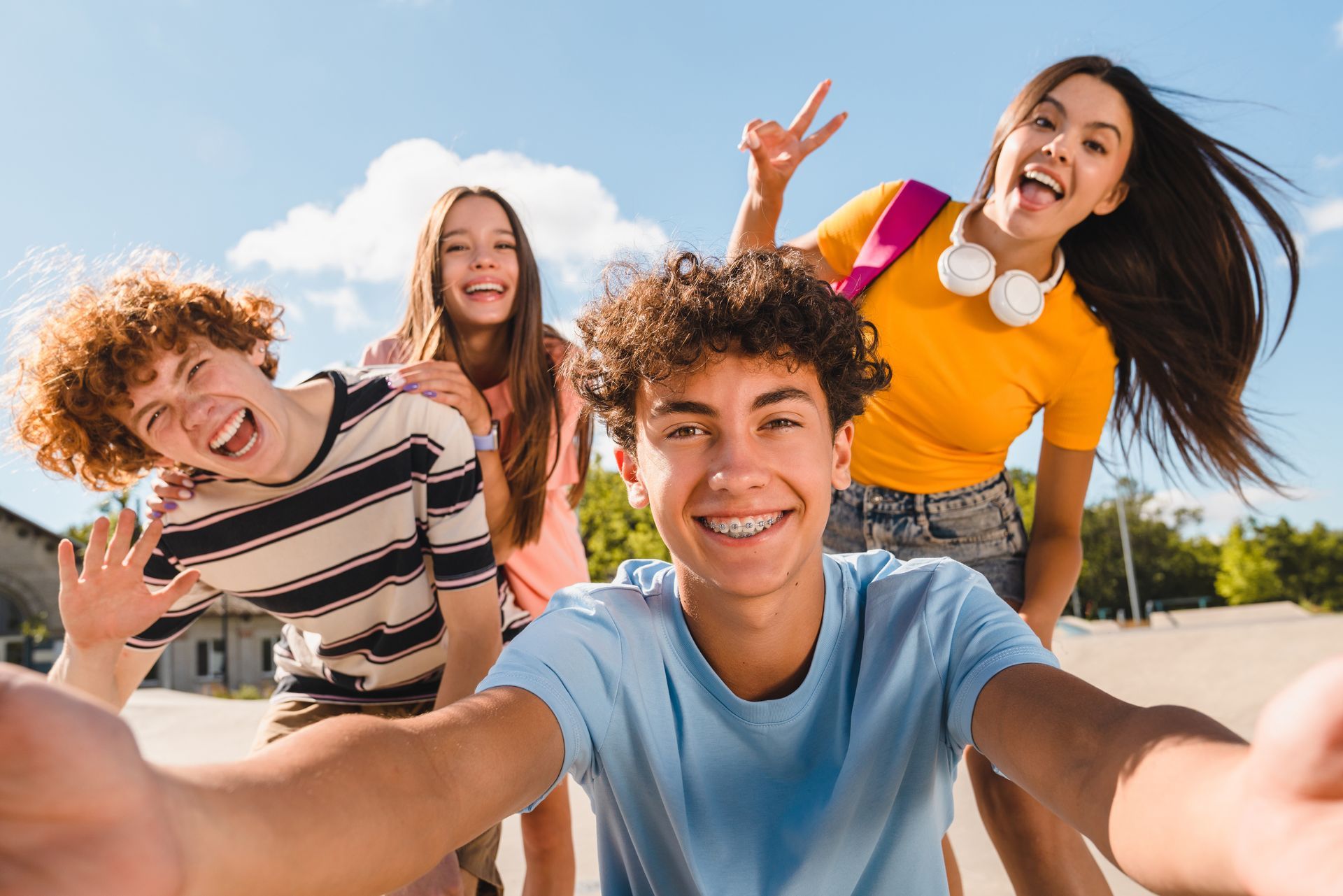 Group of teens smiling and taking a selfie outdoors on a sunny day; one person is making a peace sign.