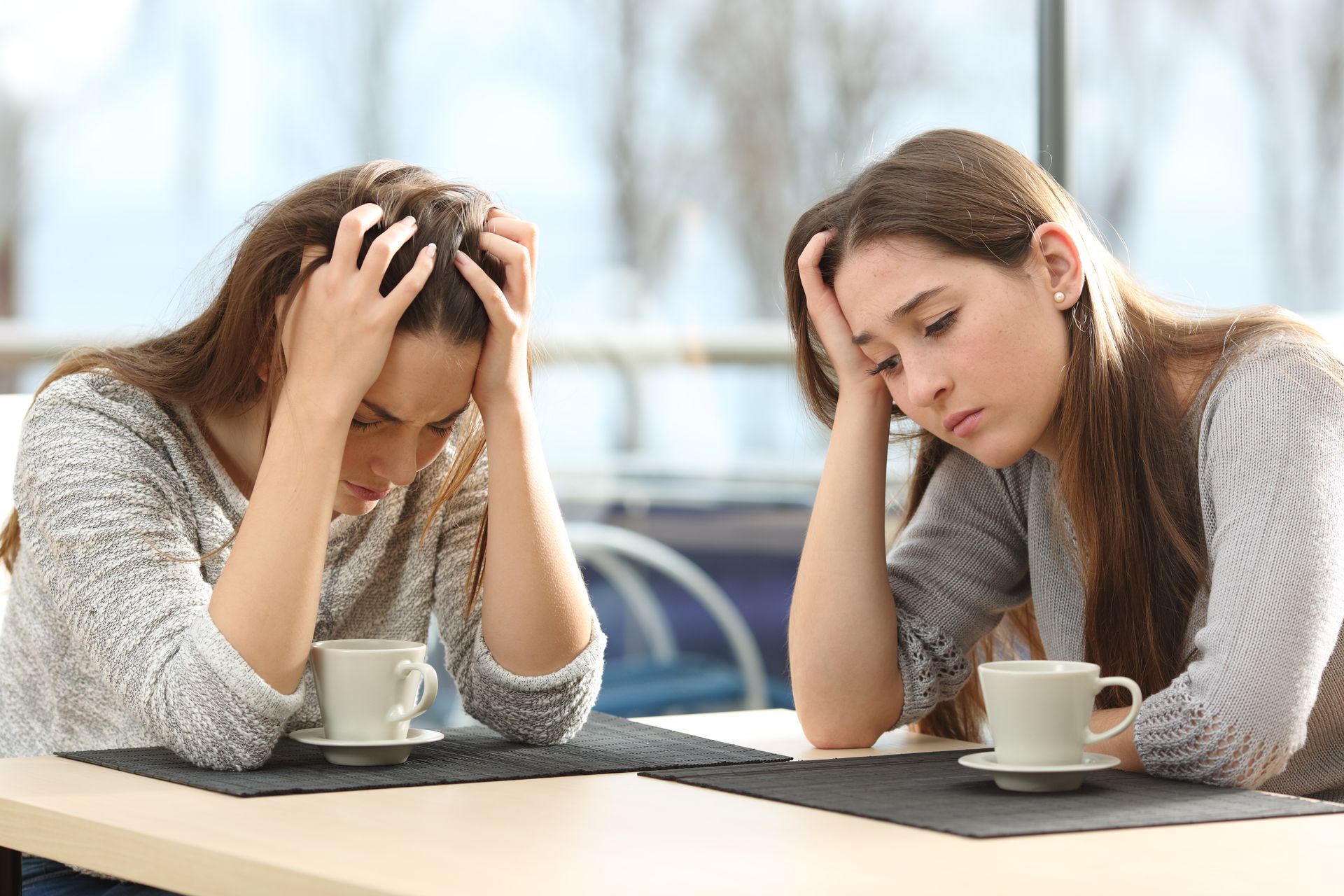 Two young women with hands on their heads, looking sad, sitting at a table in a cafe.