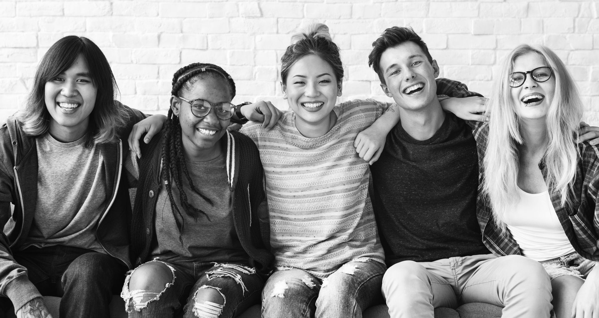 Group of five smiling young adults with arms around each other, sitting in front of a brick wall.