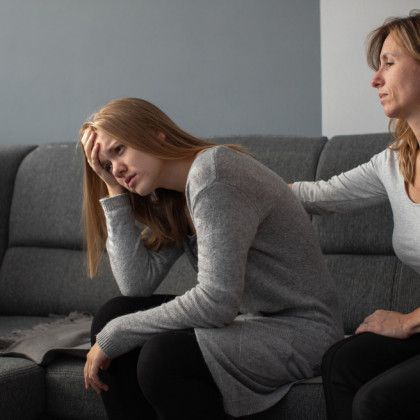 A young woman with her head in her hand, looking distressed, with a supportive woman's hand on her shoulder, sitting on a sofa.