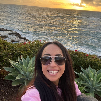 Woman with sunglasses smiles by the ocean at sunset.