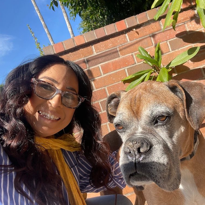 Woman with glasses smiles next to a brown boxer dog on a sunny day near a brick wall.