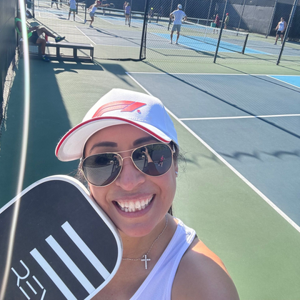 Woman smiles on pickleball court, holding a paddle. Other players visible on sunny day.
