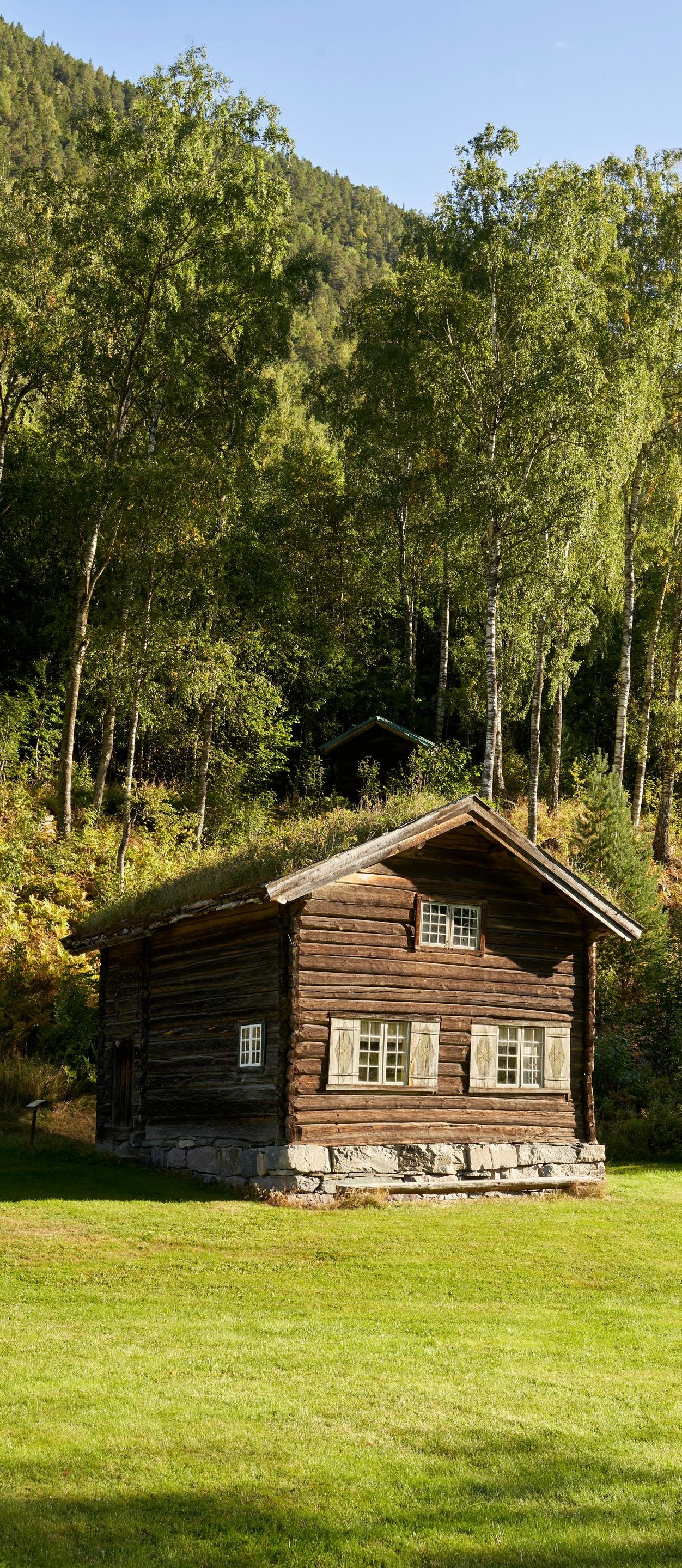 A small, dark wooden cabin with a grass roof sits on a green lawn in front of a forest and mountain slope.