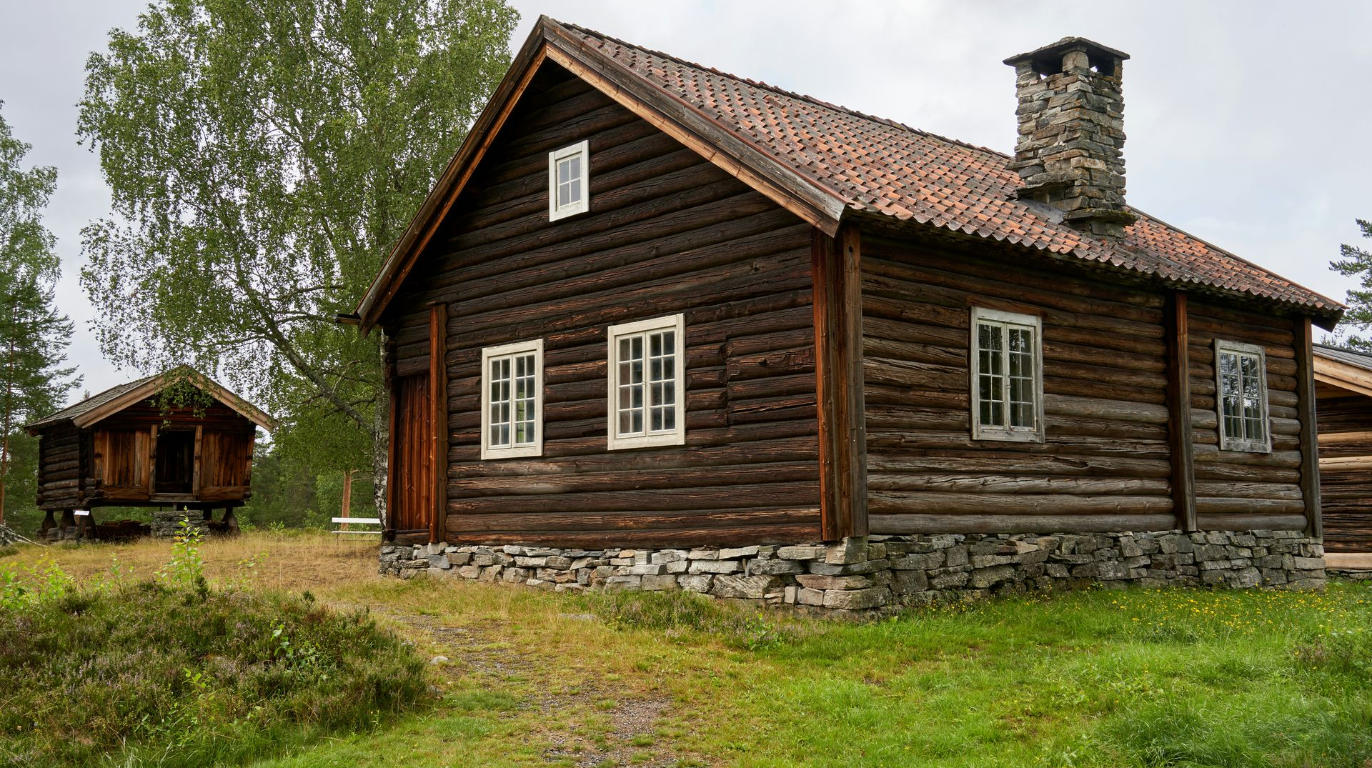 A brown log cabin with a stone foundation and chimney, surrounded by grass and trees under a cloudy sky.